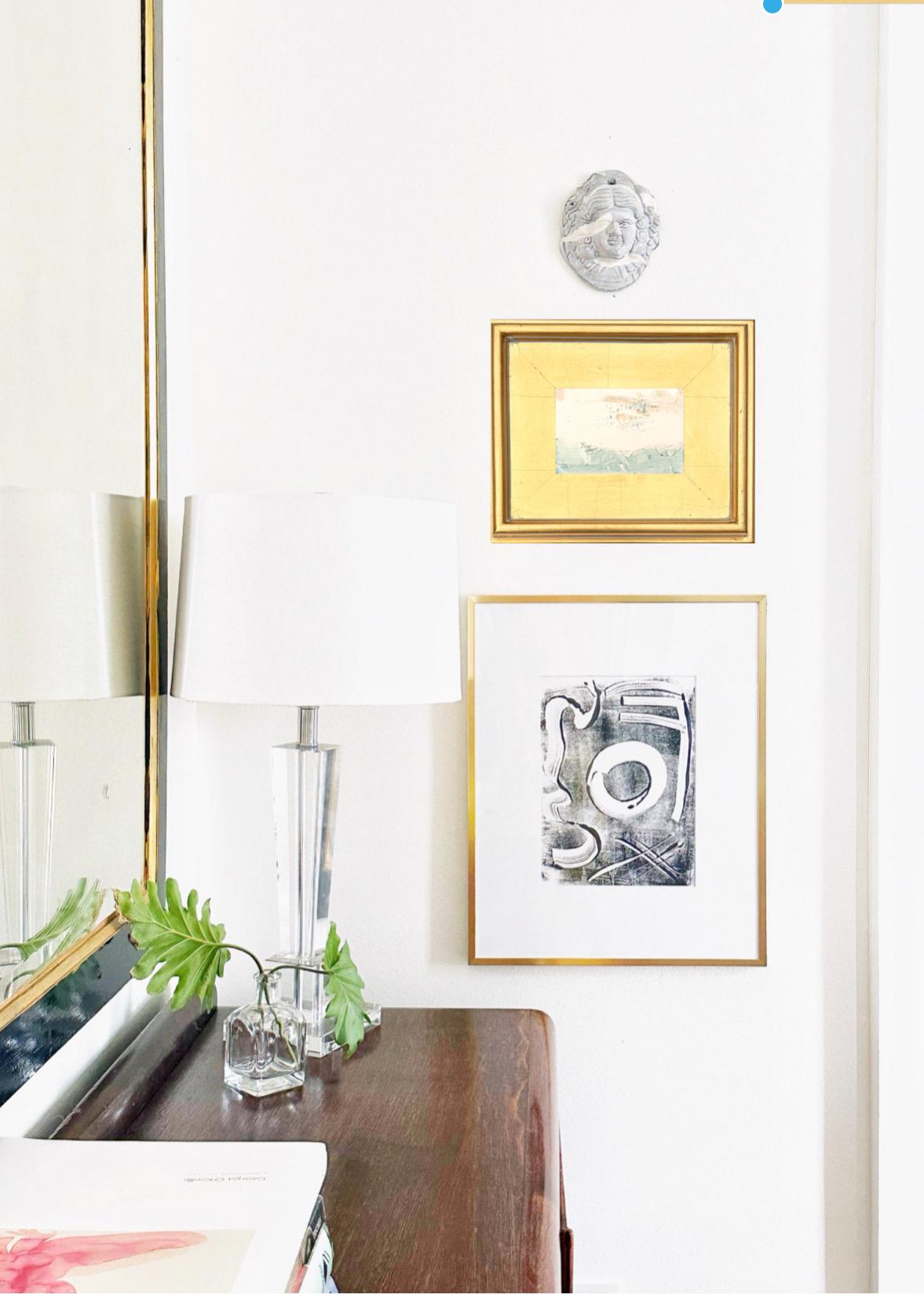 Framed original abstract artwork mixed media landscape painting in gold leaf frame with pink, light blue, and chartreuse colors on white wall above wooden dresser with lamp and greenery.