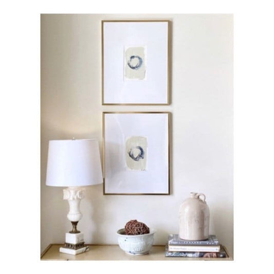 Pair of framed abstract watercolor paintings in black, white, and taupe with gold metal gallery frames, displayed above a beige table with decor.