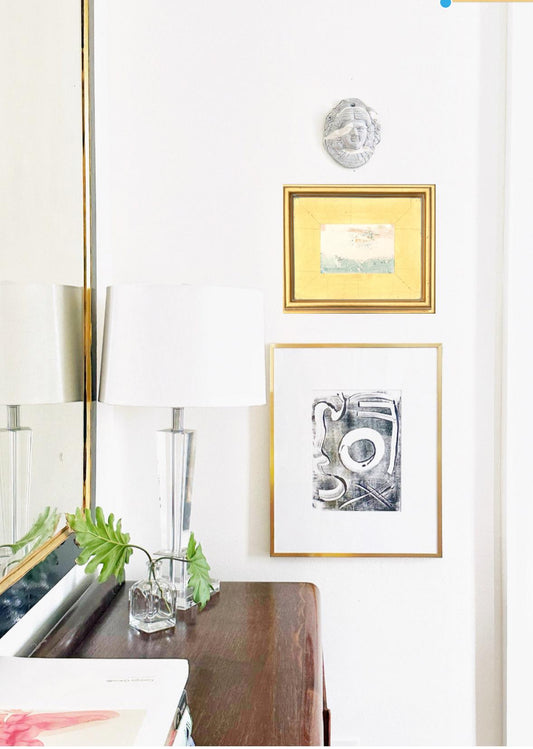 Framed original abstract artwork mixed media landscape painting in gold leaf frame with pink, light blue, and chartreuse colors on white wall above wooden dresser with lamp and greenery.